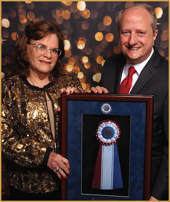 Susan Scholar holds up an award plaque/picture frame alongside another man as they both smile together