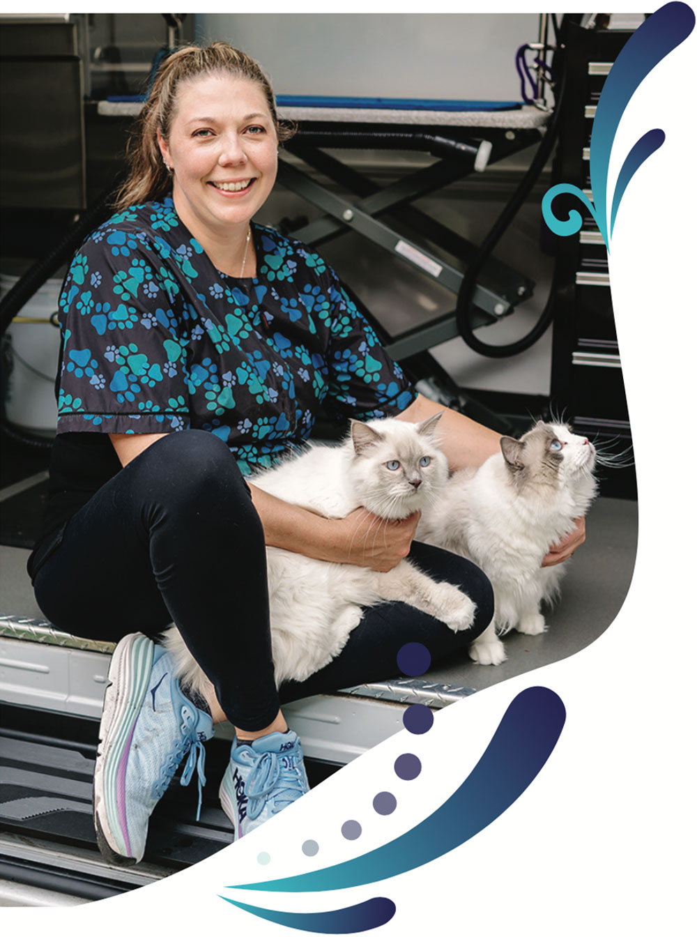 McKenna smiles at the camera while holding her hands around two white long haired blue eyed cats and sitting at the entrance of her grooming van