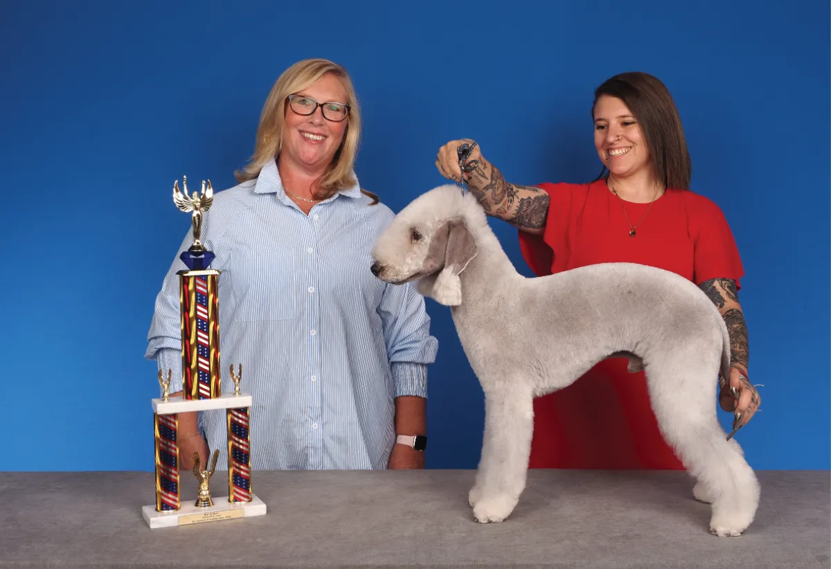 A smiling woman in a striped blue shirt standing beside a woman in a red dress holding a white Bedlington Terrier by its collar, with a large colorful trophy on the table, against a blue background.