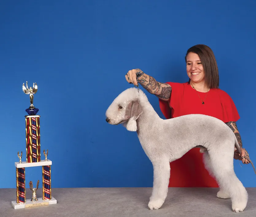 A smiling woman in a red shirt holding a white Bedlington Terrier by its collar while posing beside a large colorful trophy, against a blue background.