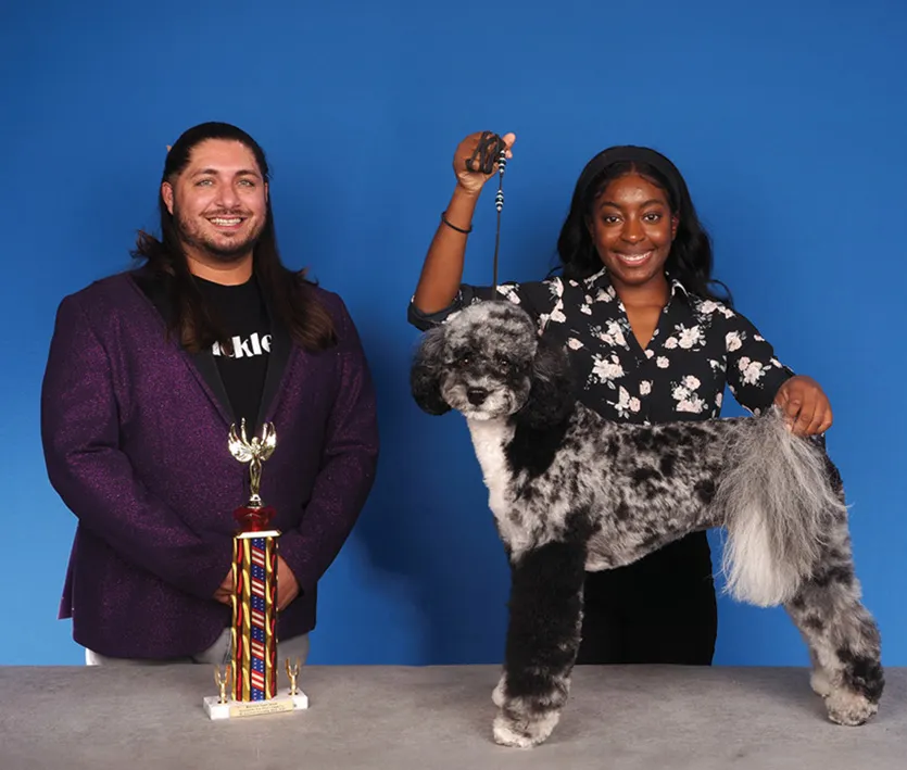 A man in a purple jacket and a woman in a floral blouse posing with a black and white poodle, with a colorful trophy on a table, against a blue background.