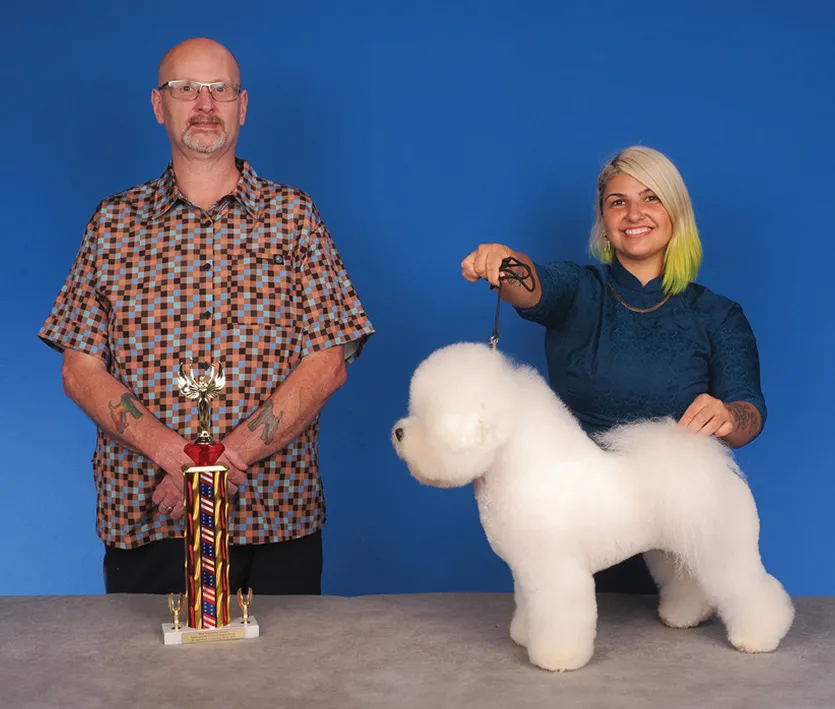 A man in a checkered shirt and a woman in a green blouse posing with a fluffy white dog, holding its collar, next to a colorful trophy, against a blue background.