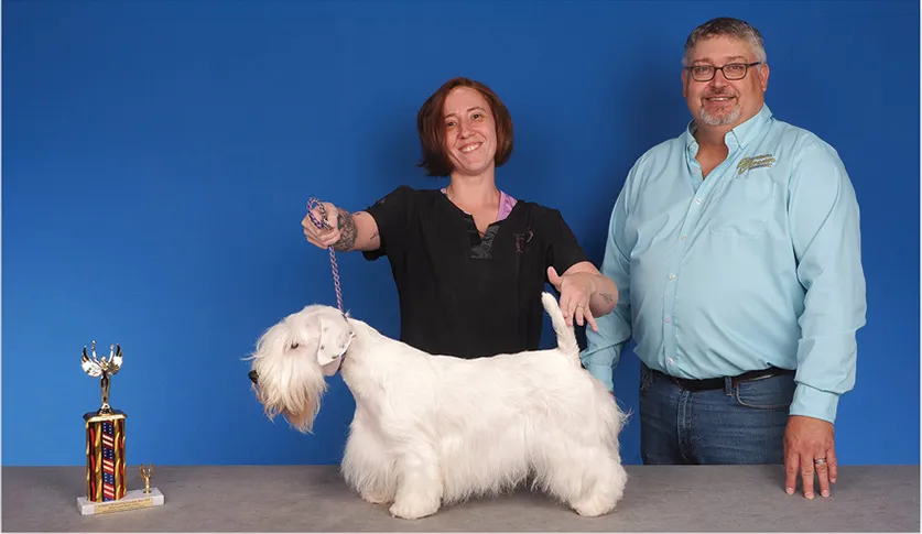 A man in a blue shirt and a woman holding a white Sealyham Terrier with a colorful trophy beside them, against a blue background.