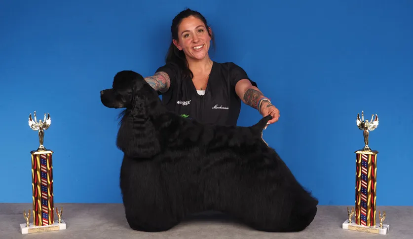 A smiling woman with tattoos in a black uniform holding a black Cocker Spaniel by its tail, with two colorful trophies on a table, against a blue background.