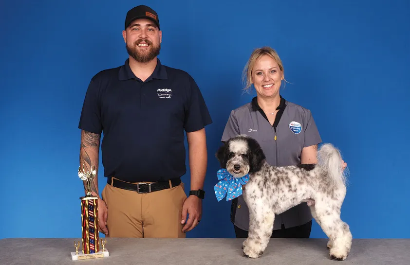 A man in a dark blue shirt and a woman in a gray uniform posing with a black and white dog wearing a blue bow, with a colorful trophy on the table, against a blue background.