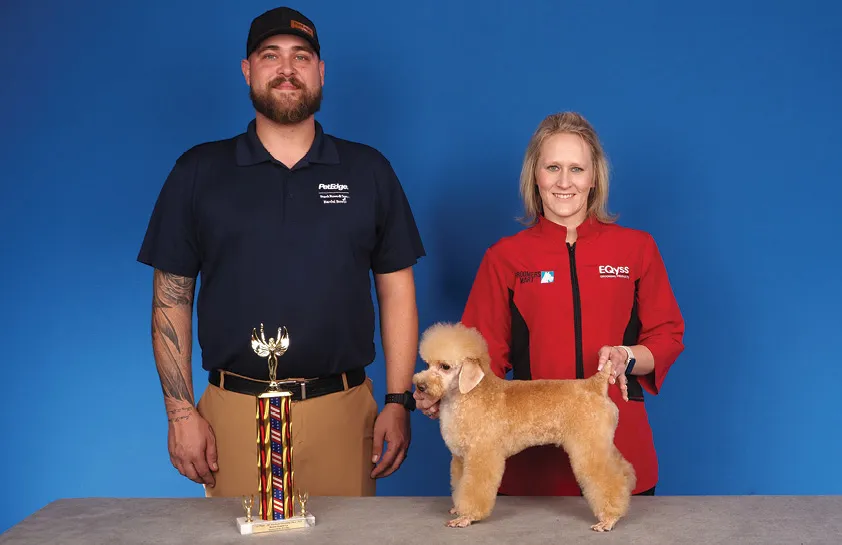 A man in a dark blue shirt and a woman in a red uniform holding a small light brown dog by its collar, with a colorful trophy on the table, against a blue background.