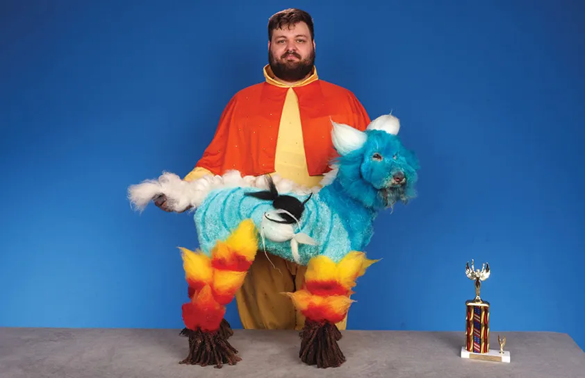 A man in an orange and yellow costume holding a creatively groomed dog with blue, white, red, and yellow fur patterns resembling a mythical creature, with a colorful trophy on the table, against a blue background.