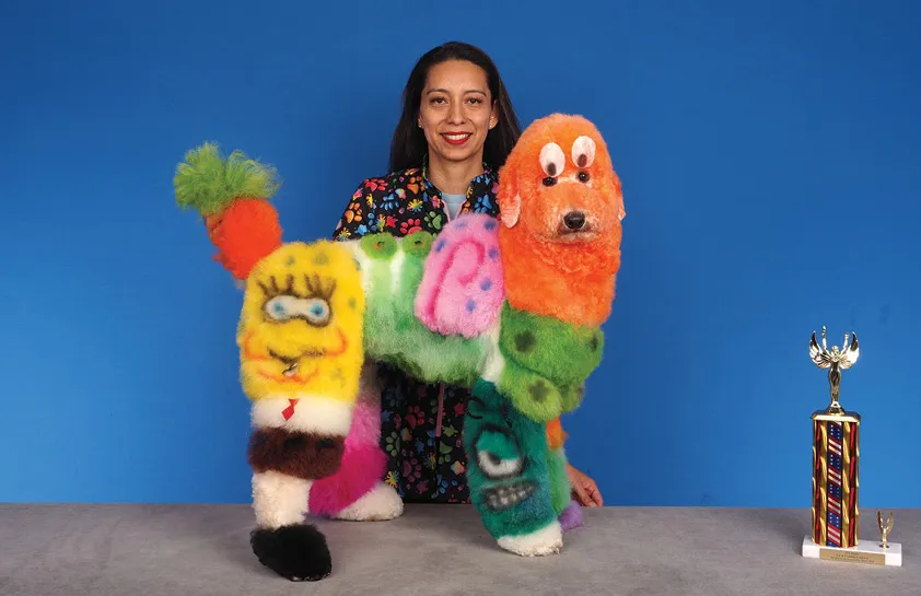 A woman in a floral shirt holding a dog groomed in a creative multicolored style, featuring cartoon character faces, with a colorful trophy on the table, against a blue background.