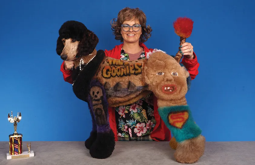 A woman in glasses, dressed in a colorful floral shirt and red jacket, holding a creatively groomed dog featuring a design inspired by 'The Goonies' with text and cartoon faces, with a colorful trophy on the table, against a blue background.