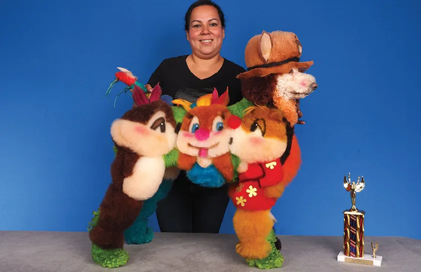 A smiling woman in a black shirt holding a creatively groomed dog with a design inspired by cartoon characters, with a colorful trophy on the table, against a blue background.