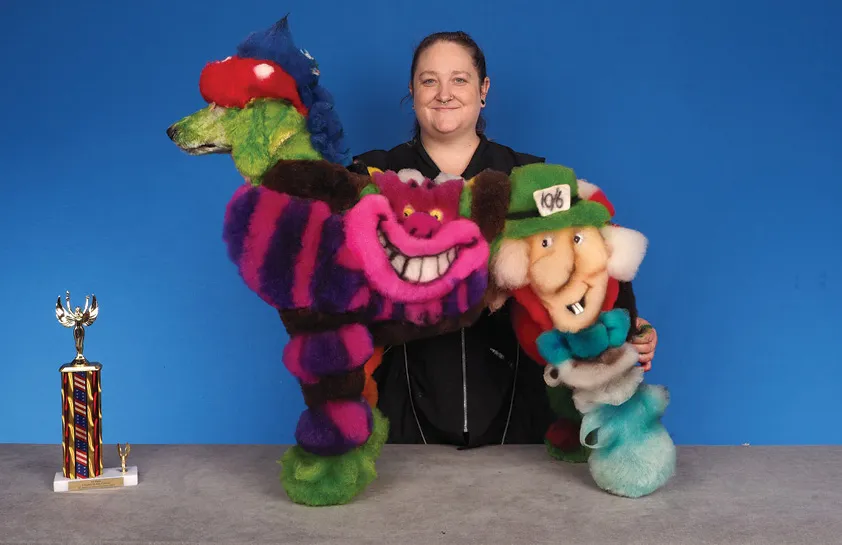 A woman in a black shirt holding a creatively groomed dog, featuring a design inspired by characters like the Cheshire Cat and a leprechaun, with a colorful trophy on the table, against a blue background.