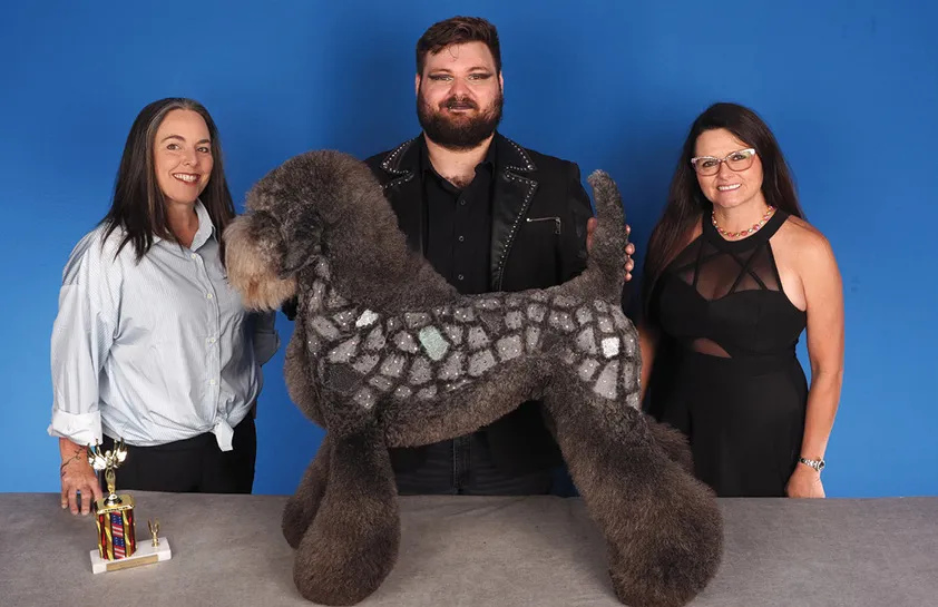 A man in a black jacket with decorative studs, flanked by two women, posing with a large poodle creatively groomed to resemble a cobblestone design, with a colorful trophy on the table, against a blue background.