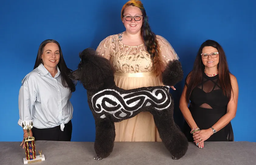 A woman in a beige gown standing between two women, holding a large black poodle groomed with intricate white Celtic knot designs on its fur, with a colorful trophy on the table, against a blue background.