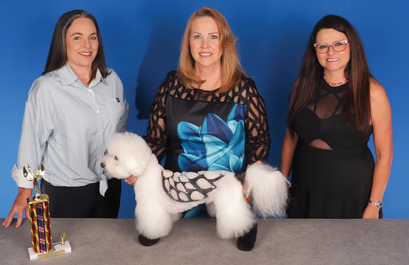 A woman in a blue floral-patterned blouse standing between two women, holding a white fluffy dog groomed with a creative black and silver pattern on its fur, with a colorful trophy on the table, against a blue background.