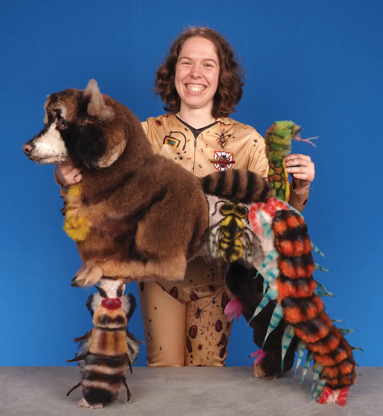 A smiling woman in a beige costume with insect prints holding a creatively groomed dog, featuring elaborate designs including a snake, insects, and other colorful elements, against a blue background.