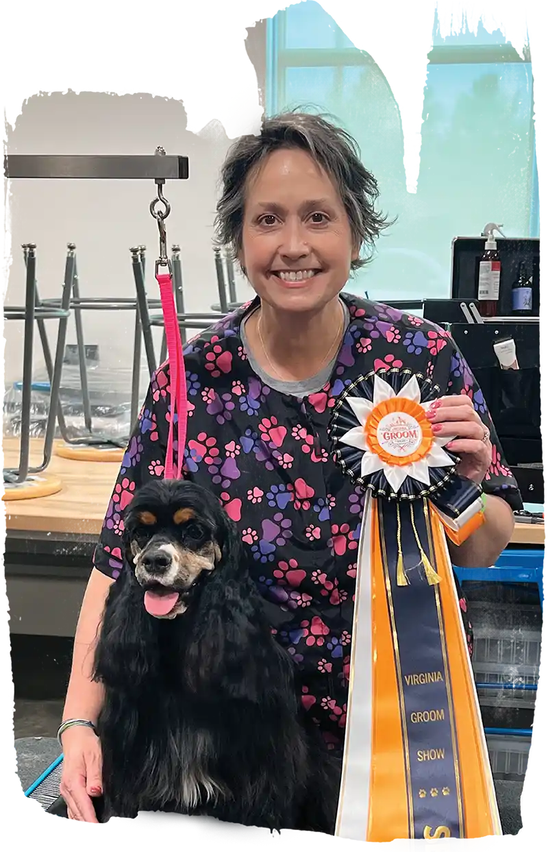 Woman smiling beside a black dog on a grooming table, holding a prize ribbon.