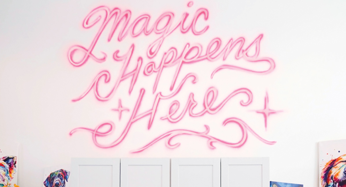 a wall with "Magic Happens Here" written in pink script, with colorful dog portraits visible below