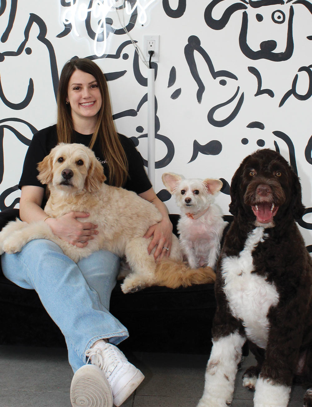 Rachel Ferreira sitting on a bench holds a cream-colored dog, while a small white dog sits beside her and a large black and white dog sits on the floor next to them, all in front of a wall with black dog-themed doodles