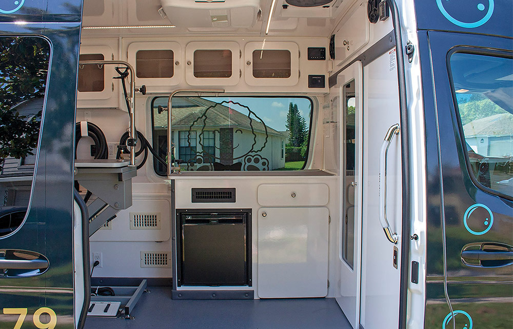 The interior of a mobile dog grooming van, showing a grooming station, cabinets, and a window with a house visible outside.