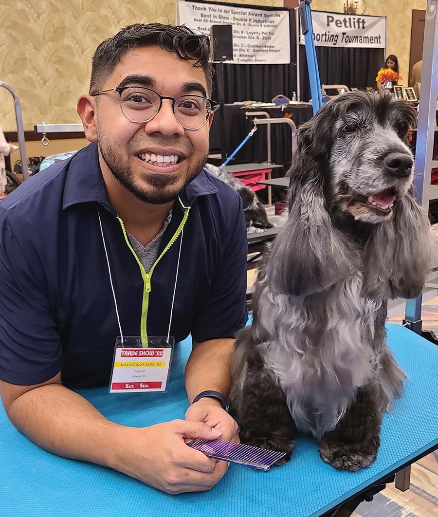 Jessie Colon-Sanchez, a man with glasses smiling and leaning beside a grey and black Cocker Spaniel on a blue grooming table in a grooming contest setting.