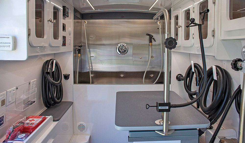 The interior of the Pup Luxe Grooming & Pet Spa mobile dog grooming van, showing a stainless steel bathing tub, a grooming table, and white cabinets.