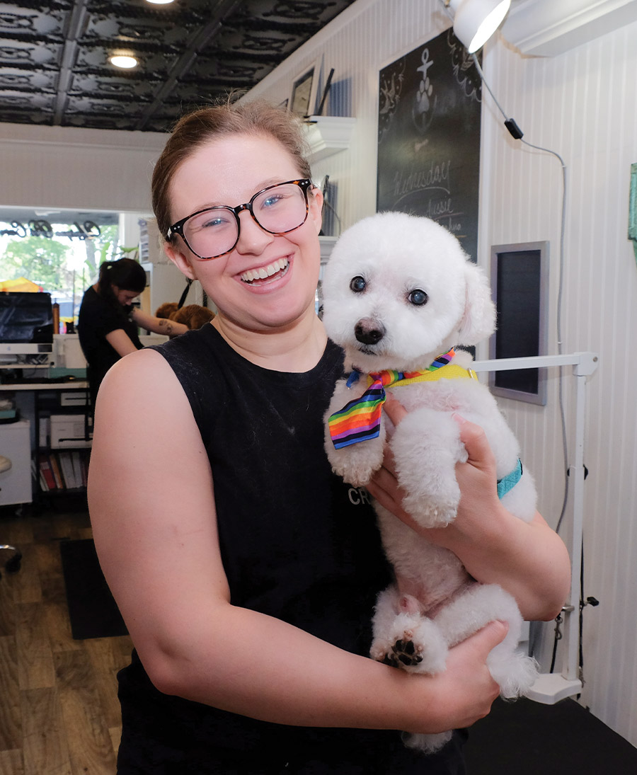 A smiling female dog groomer wearing glasses holds a small, white, fluffy dog. The dog wears a rainbow neck-tie and looks straight ahead. The woman is inside the grooming salon.