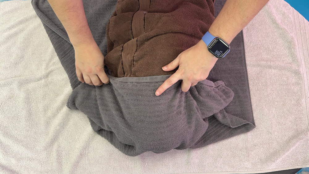 A person's hands are shown, with one hand holding and folding a gray towel over a brown towel. The person is wearing a blue smart watch on their left wrist.