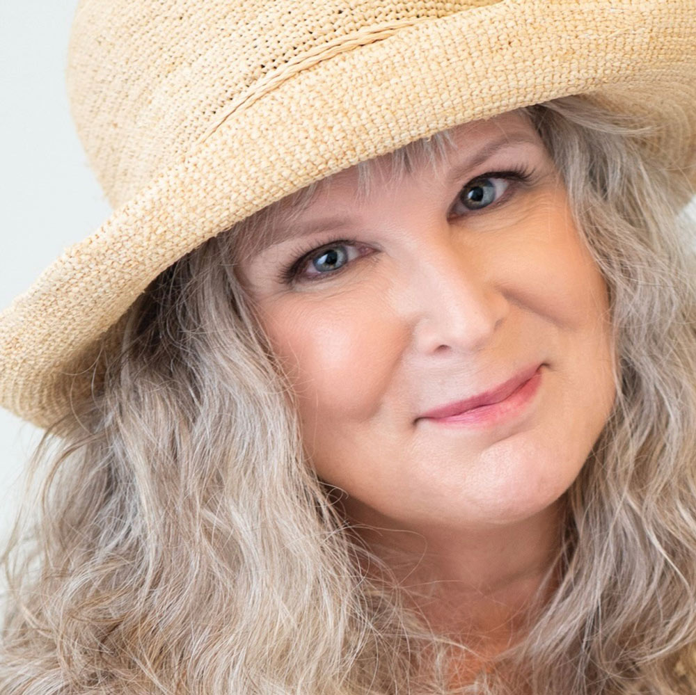 professional headshot of a smiling woman with wavy gray hair and a straw hat.