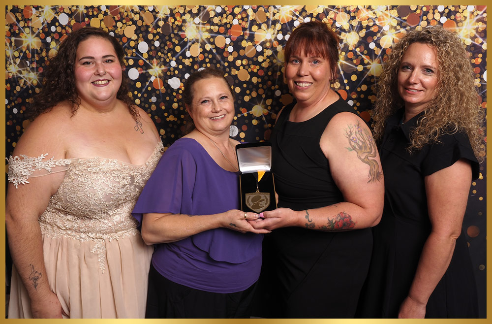 four female members of the Pet Stylist Relief Fund take a group photo as the two women in the center hold a medal presentation box displaying a metal