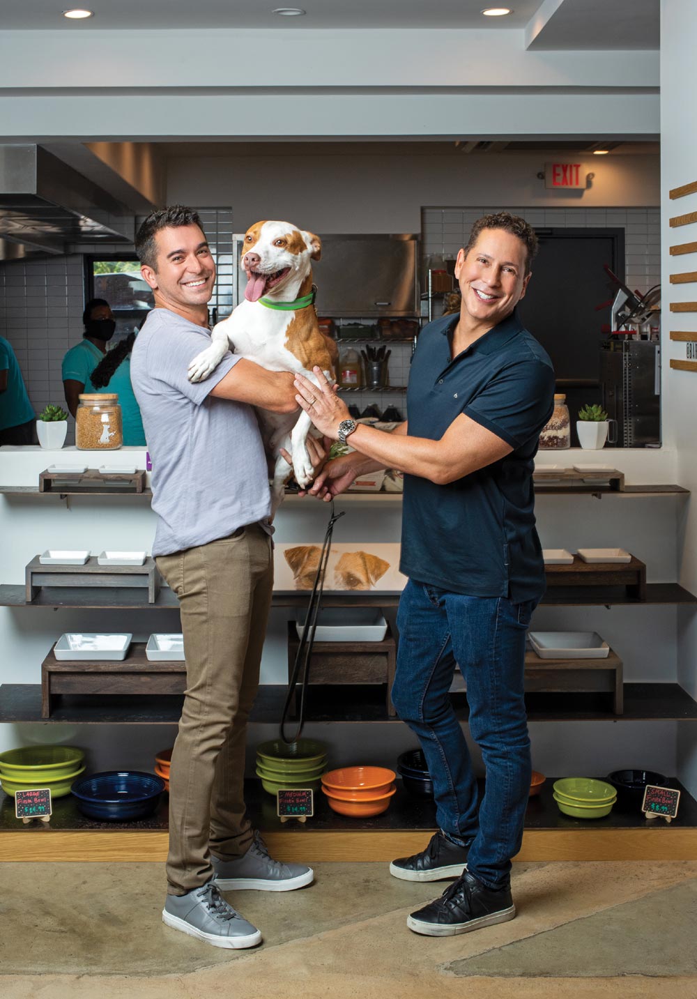 Two smiling men stand in a modern kitchen storefront; one holds a happy brown and white pit bull. Shelves with dog bowls and treats are in the background.