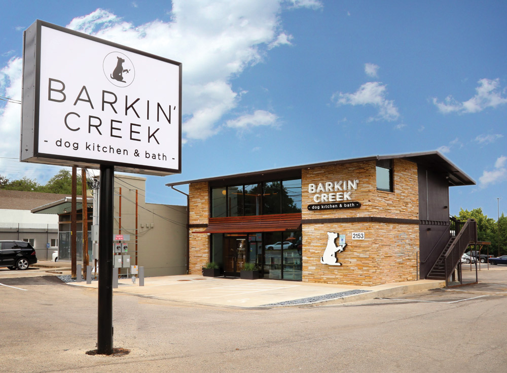 Exterior of "Barkin' Creek Dog Kitchen & Bath," a modern stone-and-glass building with a large white sign and a dog silhouette logo on the facade.