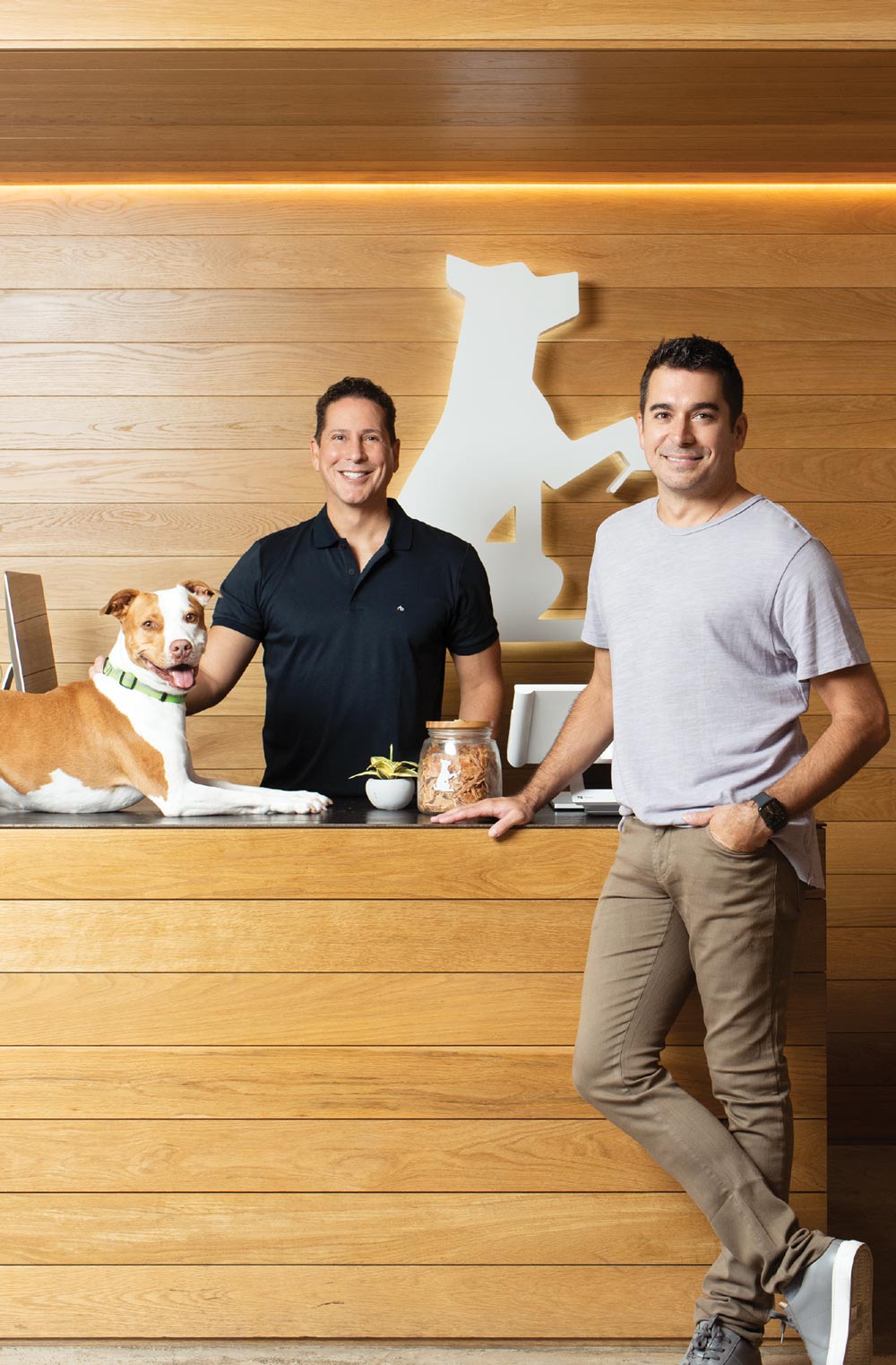 Two men, one in a black polo and one in a lilac t-shirt, smile behind a wooden reception desk. A brown and white pit bull rests on the counter next to a treat jar.