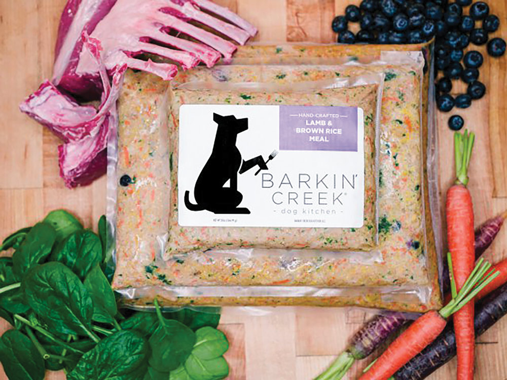 Vacuum-sealed "Lamb & Brown Rice Meal" dog food by Barkin' Creek, surrounded by fresh ingredients: raw lamb ribs, blueberries, spinach, and whole carrots.