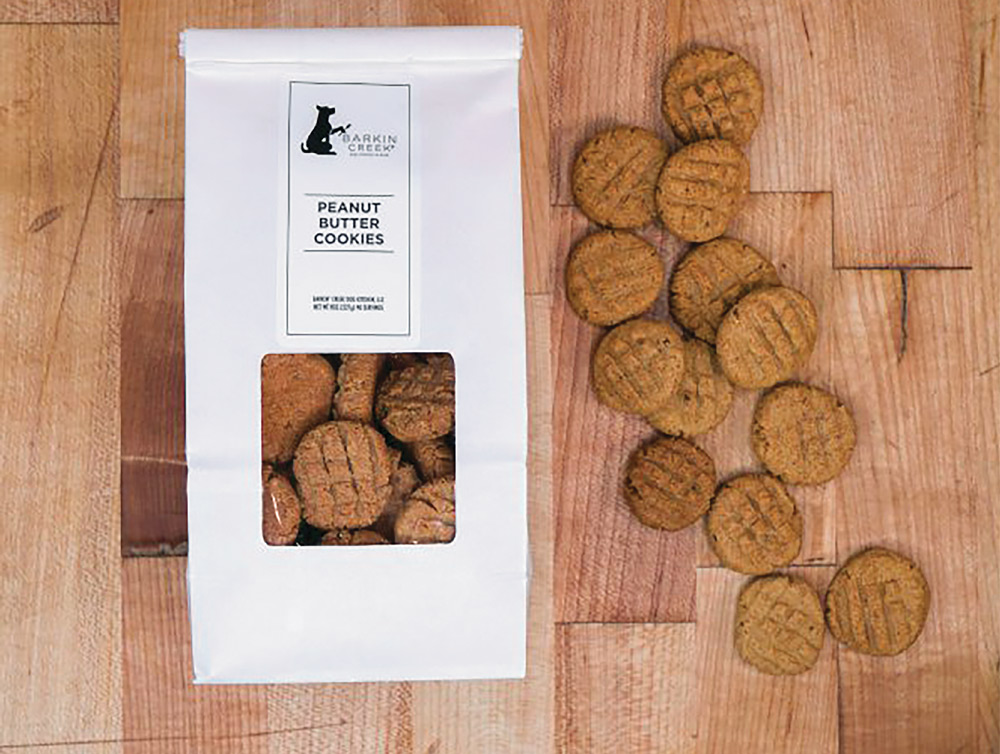 A white bag of Barkin' Creek "Peanut Butter Cookies" for dogs next to several round, cross-hatched treats scattered on a light-colored wooden cutting board.