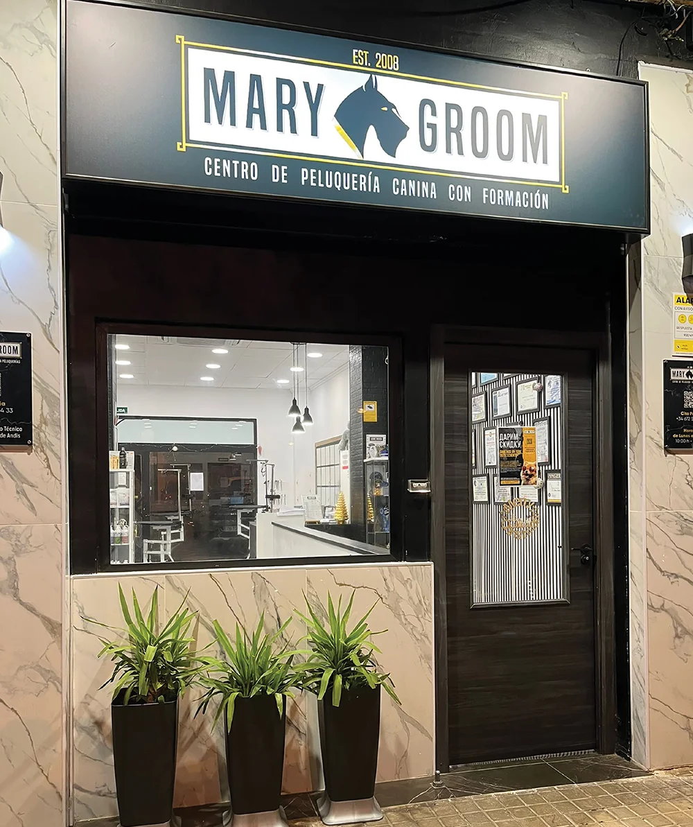 Exterior of Mary Groom, a modern dog grooming salon and training center. The dark storefront features a large sign, a glass door, and three potted plants on a marble-tiled sidewalk.