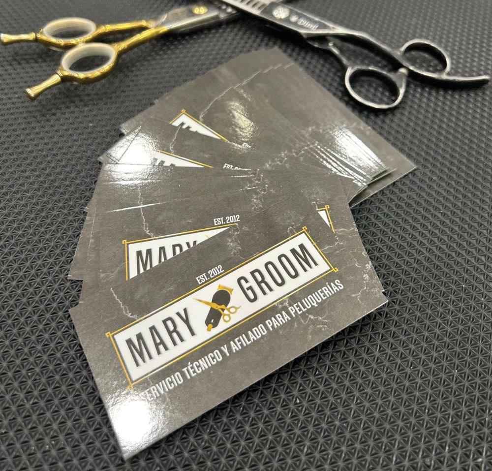 Close-up of Mary Groom business cards resting on a black textured mat next to professional grooming shears. The cards highlight technical service and sharpening for salons.