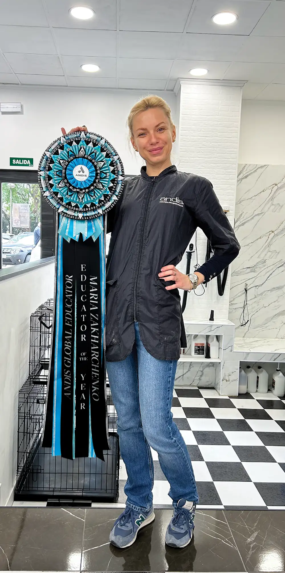 Maria Zakharchenko stands in a grooming salon holding a large, ornate blue and black award ribbon. Text on the ribbon honors her as the Andis Global Educator of the Year.
