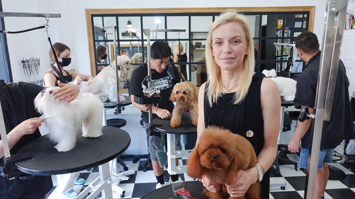 Maria Zakharchenko smiles in a busy grooming studio with a checkered floor. In the background, students at several stations work on grooming various small dogs like poodles.