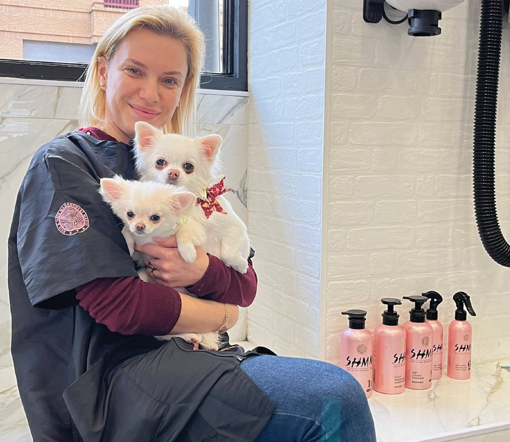 Maria Zakharchenko sits in a bright grooming room holding two small, white long-haired Chihuahuas. A row of pink SHMN brand grooming spray bottles is lined up on the counter.