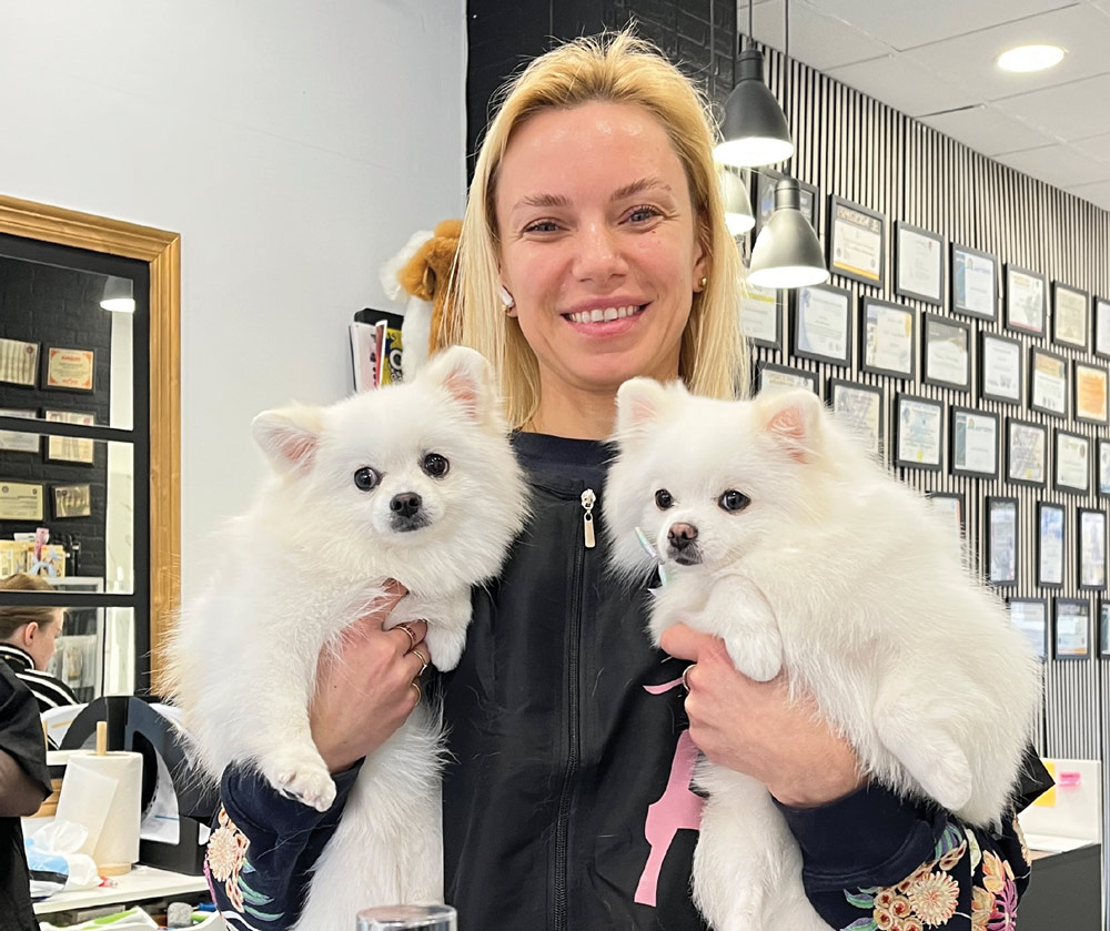 Maria Zakharchenko smiles warmly while holding two fluffy, white Pomeranian dogs. The background wall is covered in numerous framed professional certifications and awards.