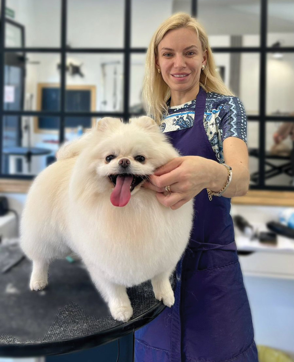 Maria Zakharchenko poses with a perfectly groomed, fluffy white Pomeranian on a grooming table. She wears a purple apron in a bright studio with a large windowed partition.