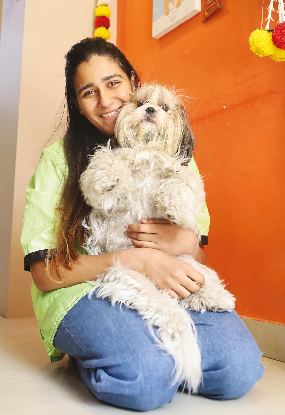 A smiling woman in a lime green grooming shirt sits on the floor holding a fluffy, freshly groomed white and tan Shih Tzu dog.