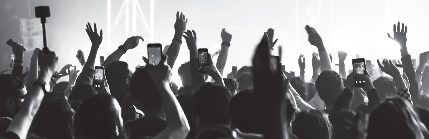 A black and white photograph of a dense, silhouetted concert crowd with a multitude of hands and illuminated smartphone screens raised high in the air, recording the bright stage.