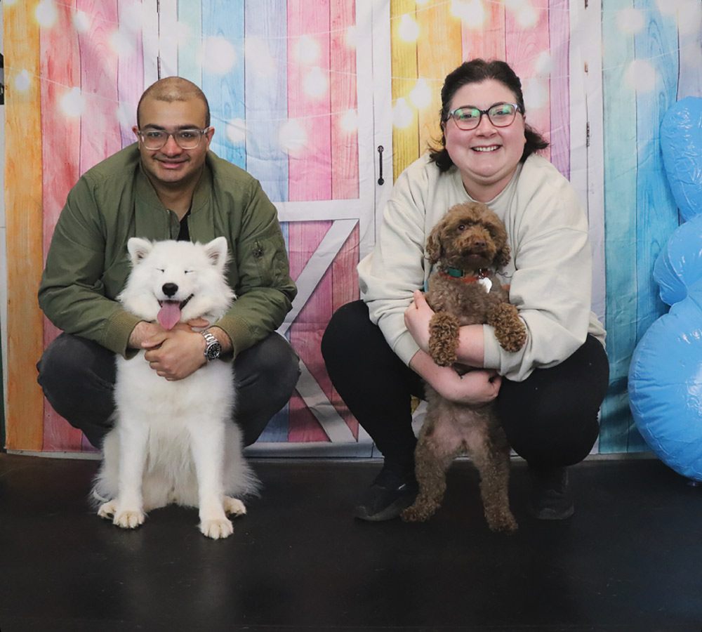 A man and a woman squatting in front of a colorful wooden backdrop, each holding a small dog.
