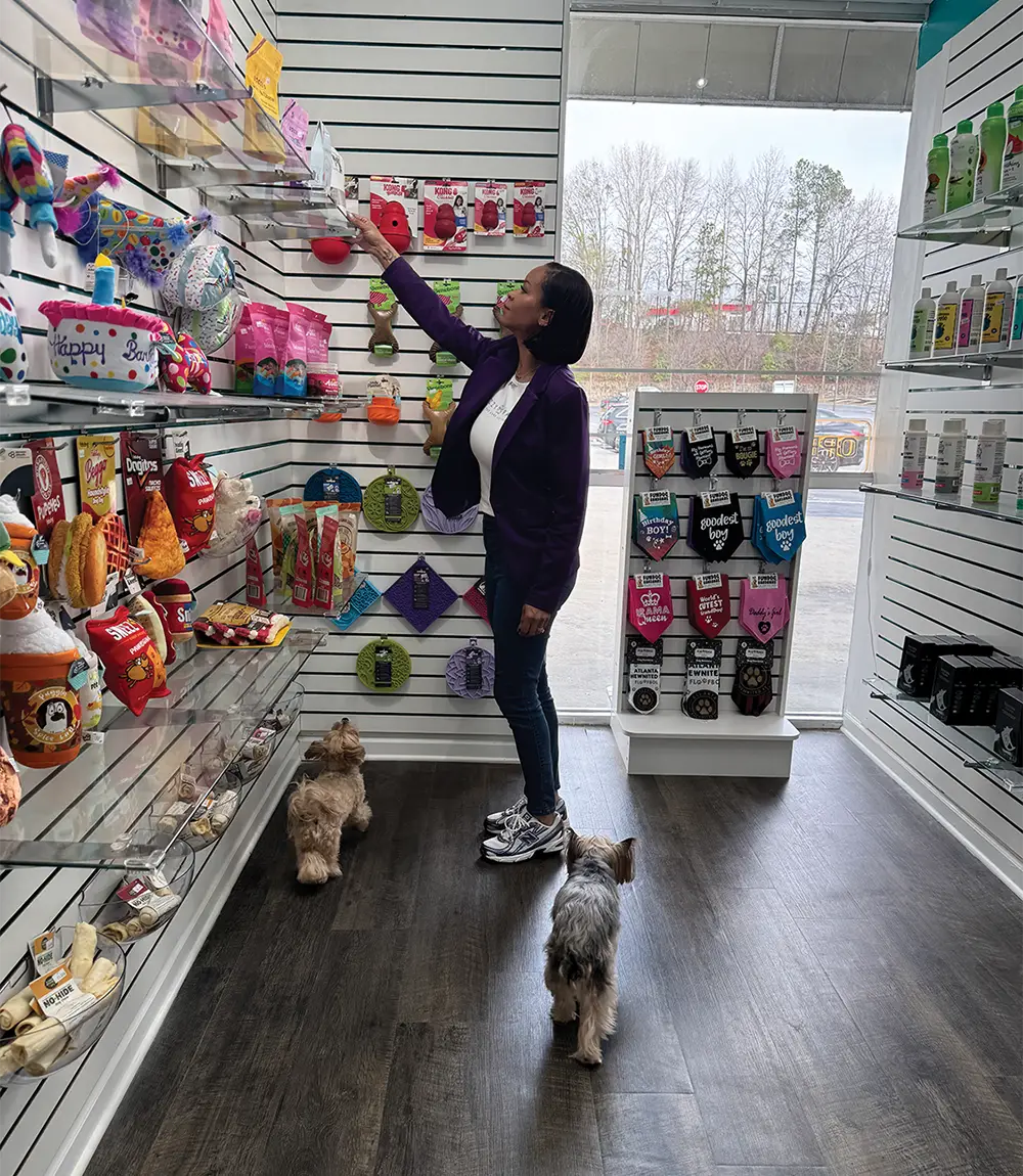 Andrea Jenkins reaches for an item on a pet product display wall while two small dogs watch from the floor.