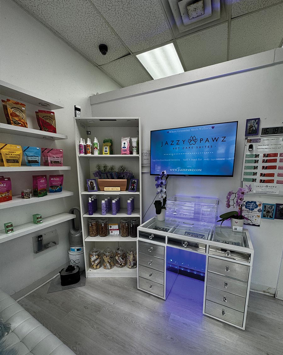A salon reception area with shelves of pet products and a silver vanity desk under a television screen displaying the Jazzy Pawz logo.