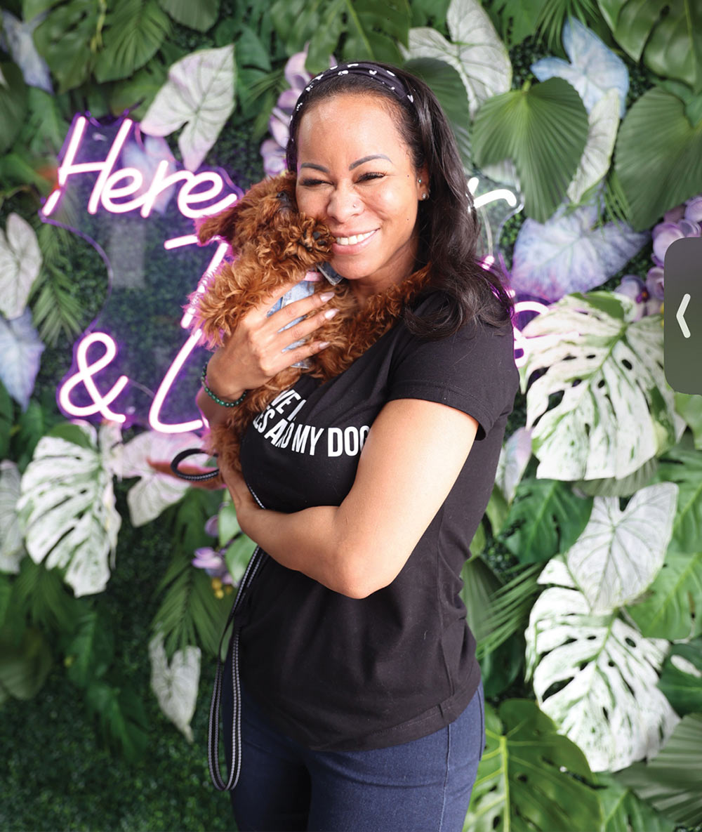 Andrea Jenkins smiles while hugging a small, brown, fluffy puppy in front of a green leaf wall with a purple neon sign.