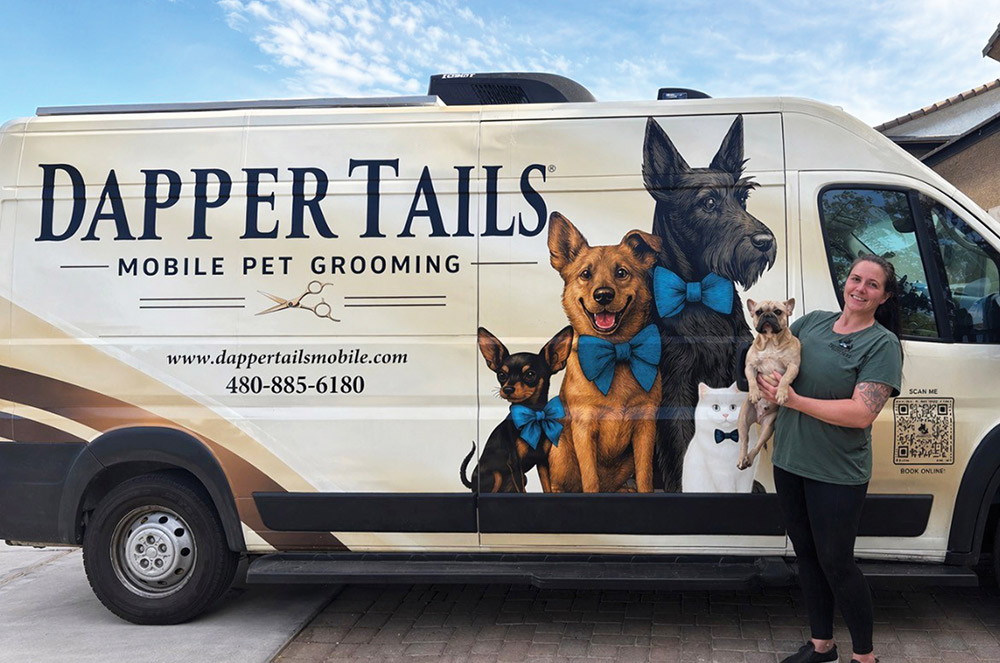 A woman holds a small dog while standing next to a white Dapper Tails Mobile Pet Grooming van. The van features a large graphic of three dogs and a white cat, all wearing blue bow ties.