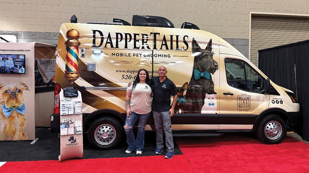 A woman and a man stand together in front of a Dapper Tails Mobile Pet Grooming van on a red carpet at an indoor event. The van features a graphic of pets wearing blue bow ties and a barber pole design.