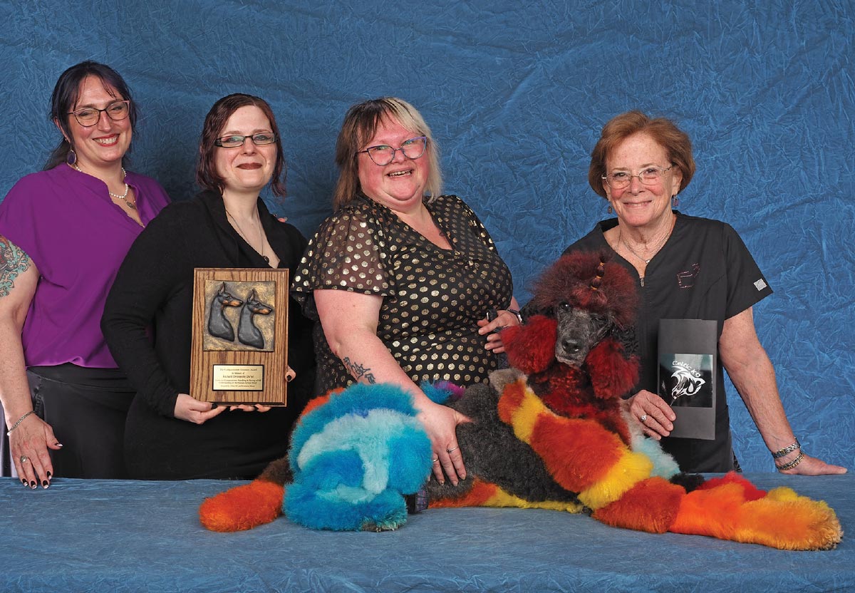 Four women posing with a plaque and a Poodle dyed in vibrant red, orange, yellow, and blue creative colors.